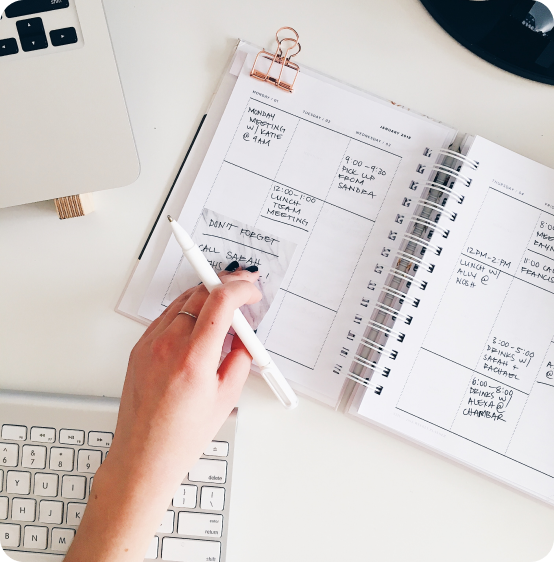 Stock image overhead shot of a hand holding a pen over a planner, a keyboard, and the corner of a laptop and mouse.